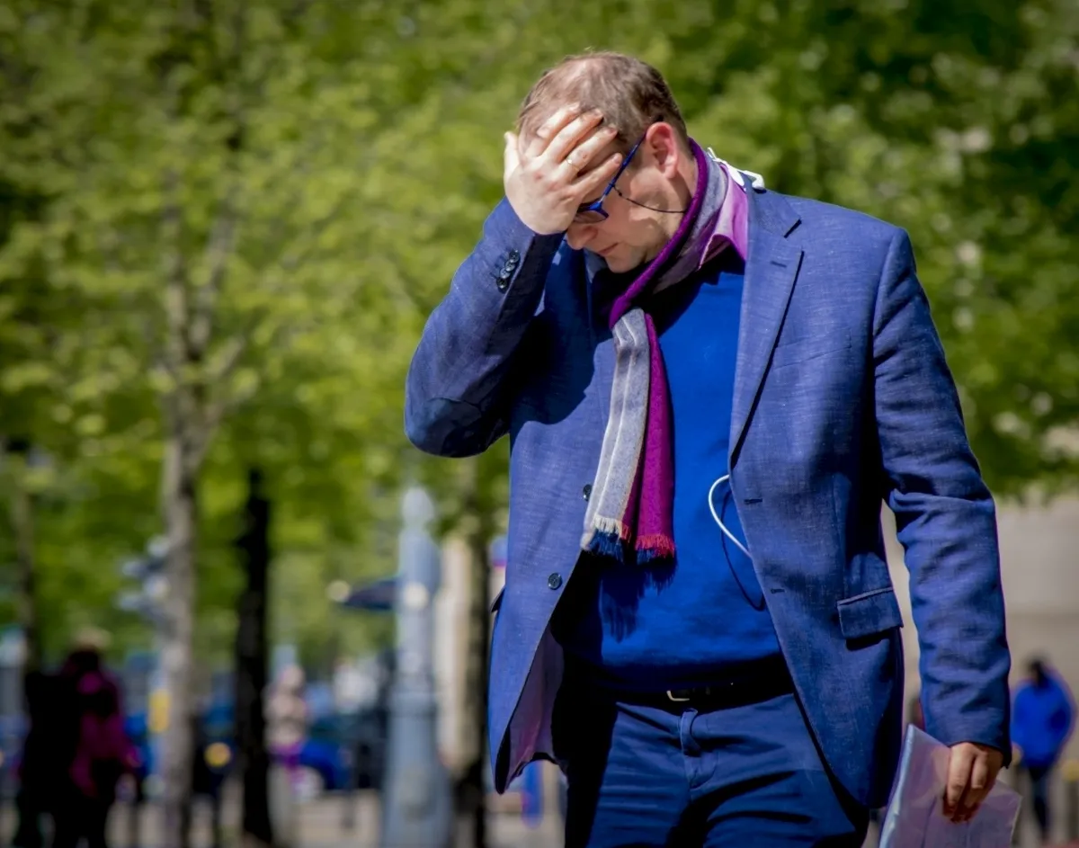 Man in a blue suit covering his forehead with one hand on a tree-lined street